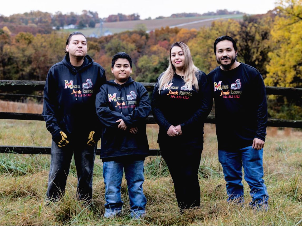 Four people pose for a photo outdoors; all wear matching black sweatshirts. A grassy field and fall trees are in the background.