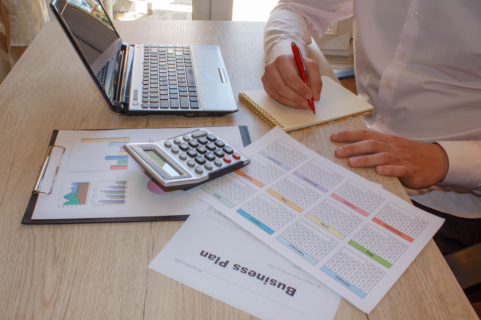 Person writing with red pen, surrounded by business documents, calculator, and laptop on a light wood desk. Person writing with red pen, surrounded by business documents, calculator, and laptop on a light wood desk.