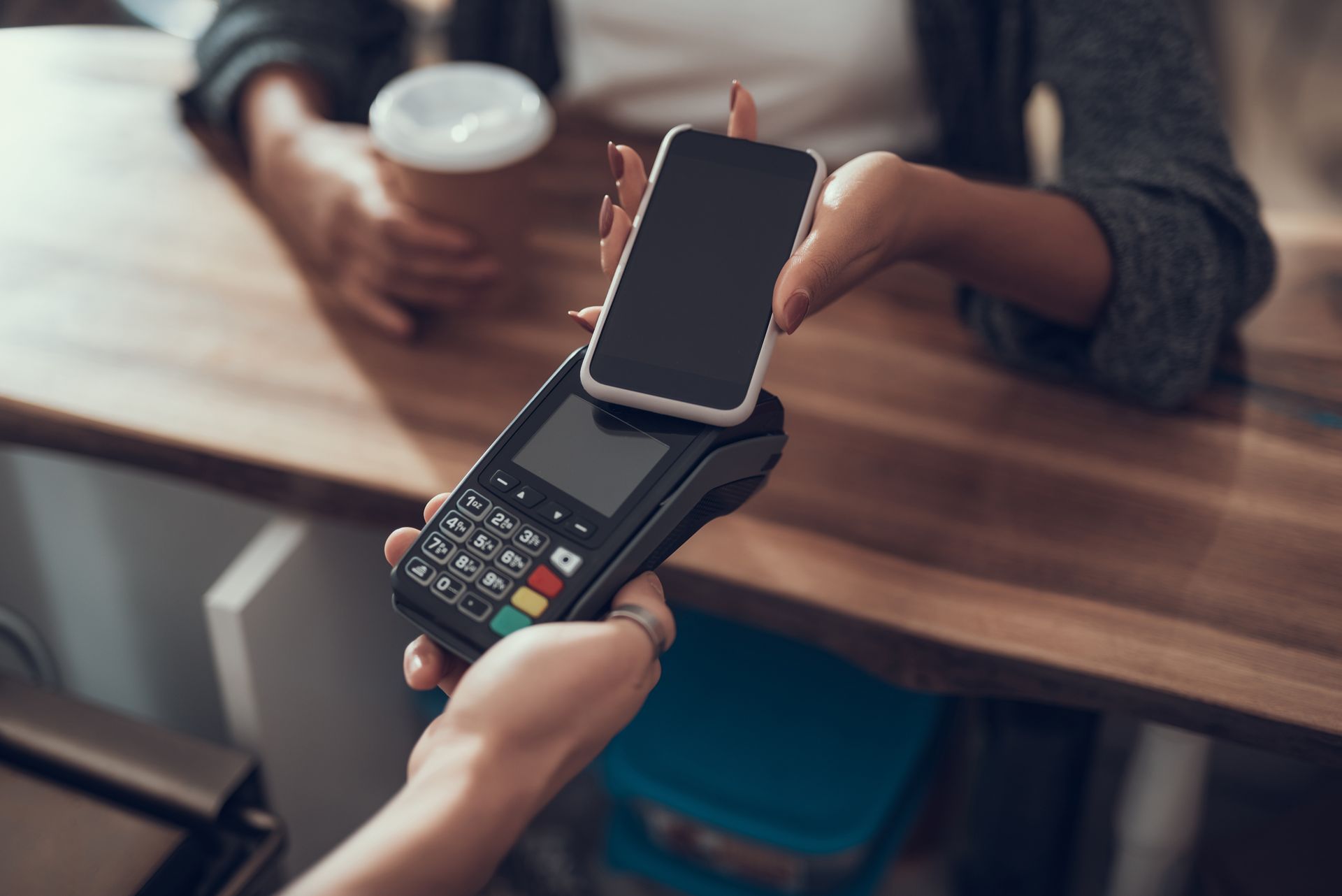 Person paying with phone at a point-of-sale terminal. A coffee cup is visible. Person paying with phone at a point-of-sale terminal. A coffee cup is visible.