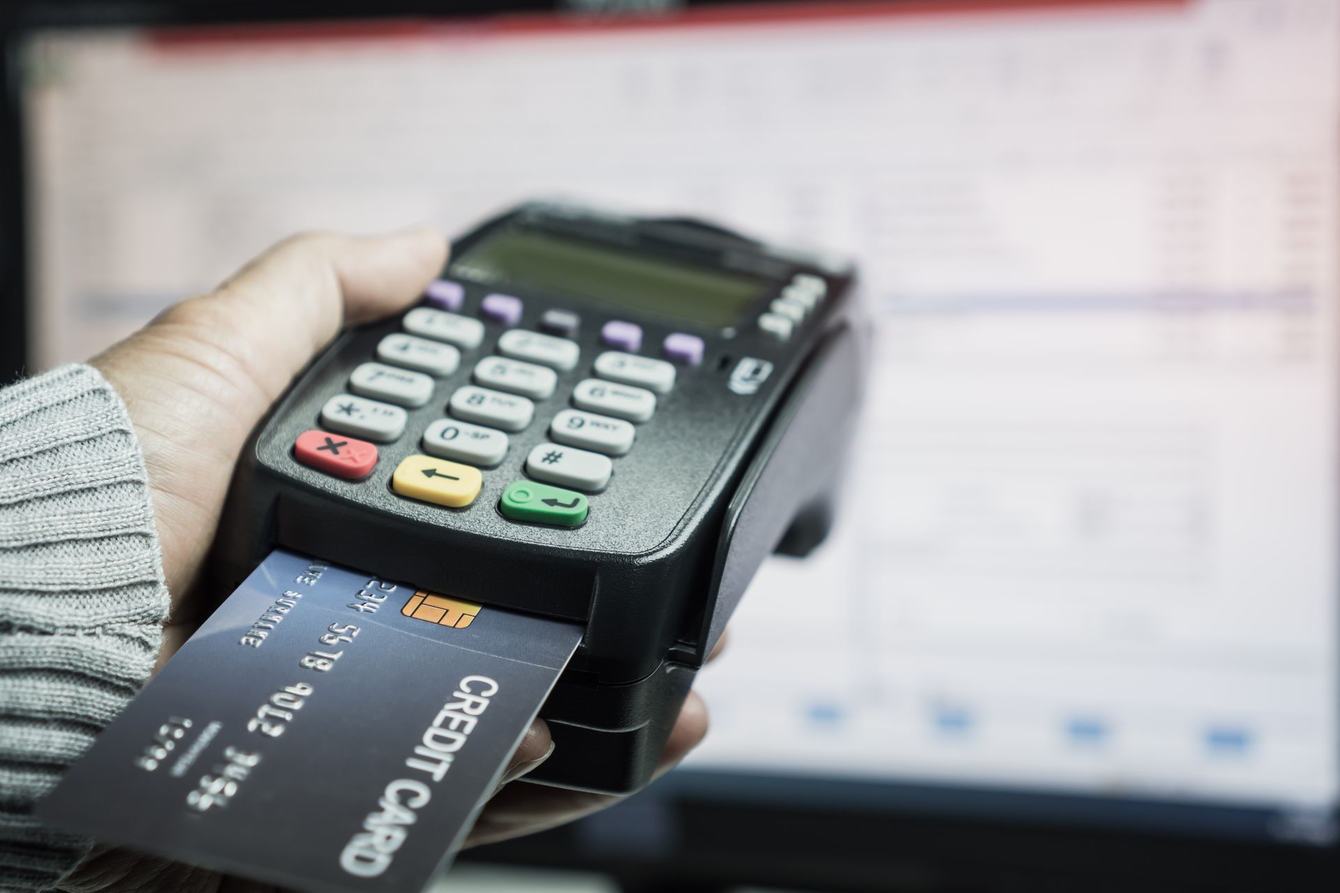 Person holding a credit card inserted into a black card reader with a computer screen in the background. Person holding a credit card inserted into a black card reader with a computer screen in the background.