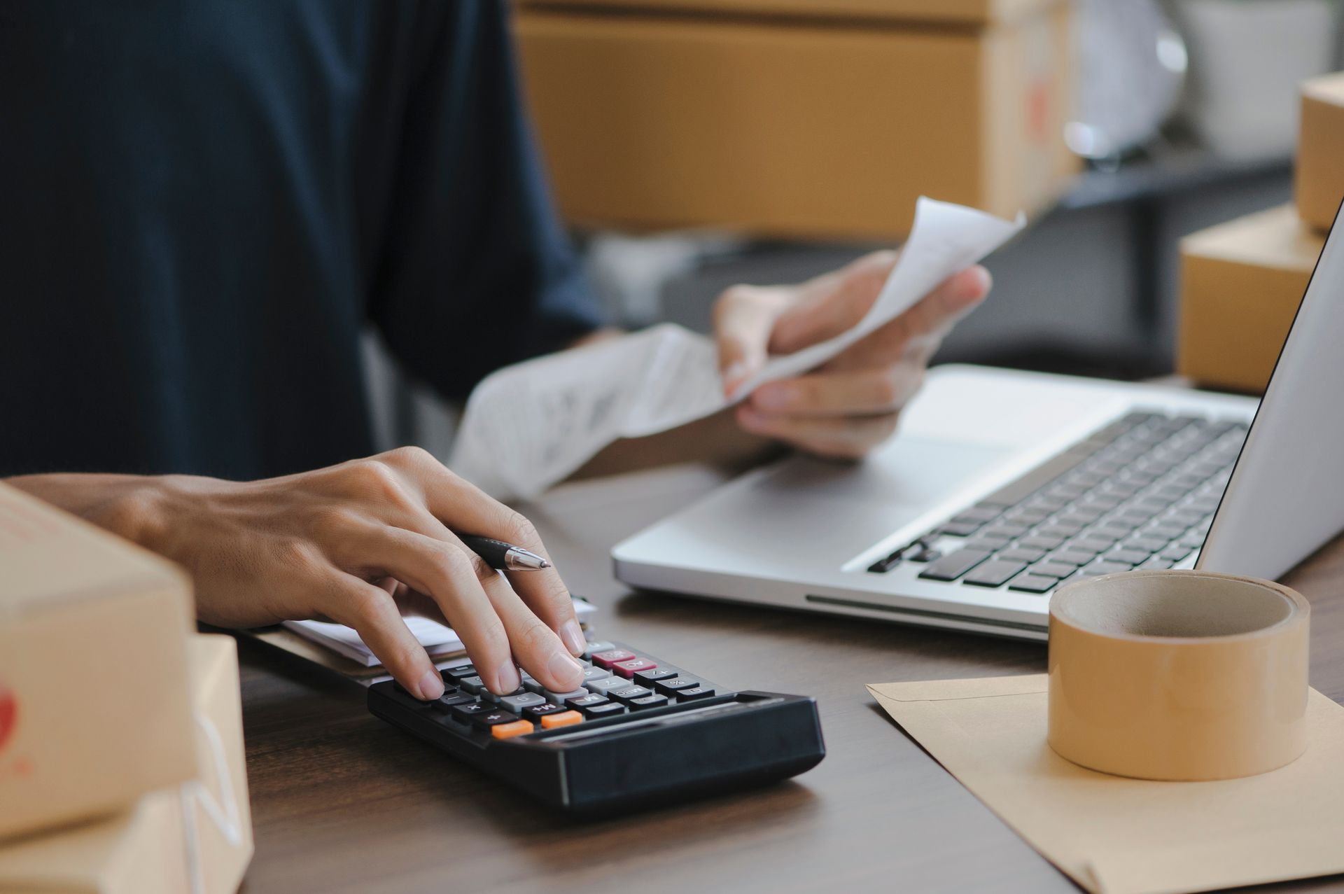 Person using a calculator, holding a receipt, and working on a laptop surrounded by shipping boxes. Person using a calculator, holding a receipt, and working on a laptop surrounded by shipping boxes.