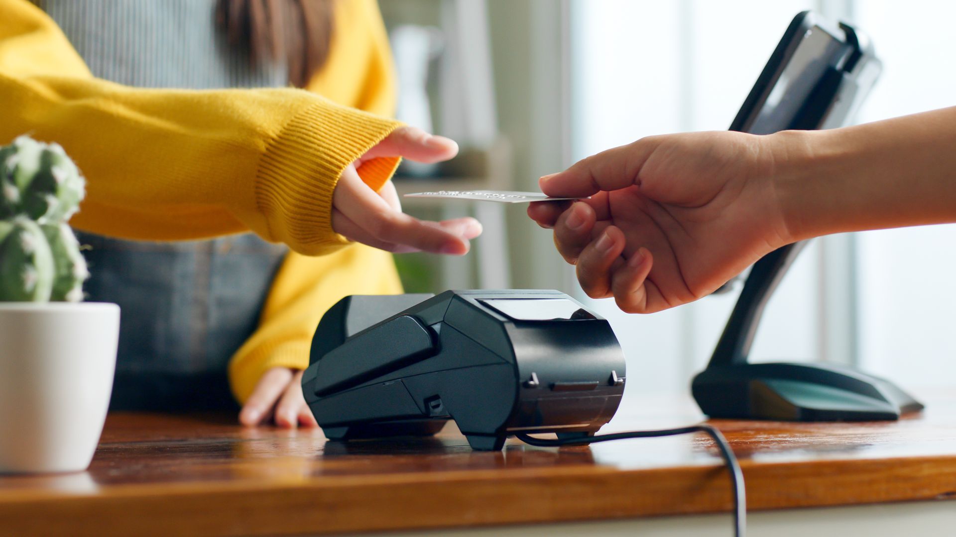 A person in a yellow sweater hands a card to a cashier near a payment terminal. A person in a yellow sweater hands a card to a cashier near a payment terminal.