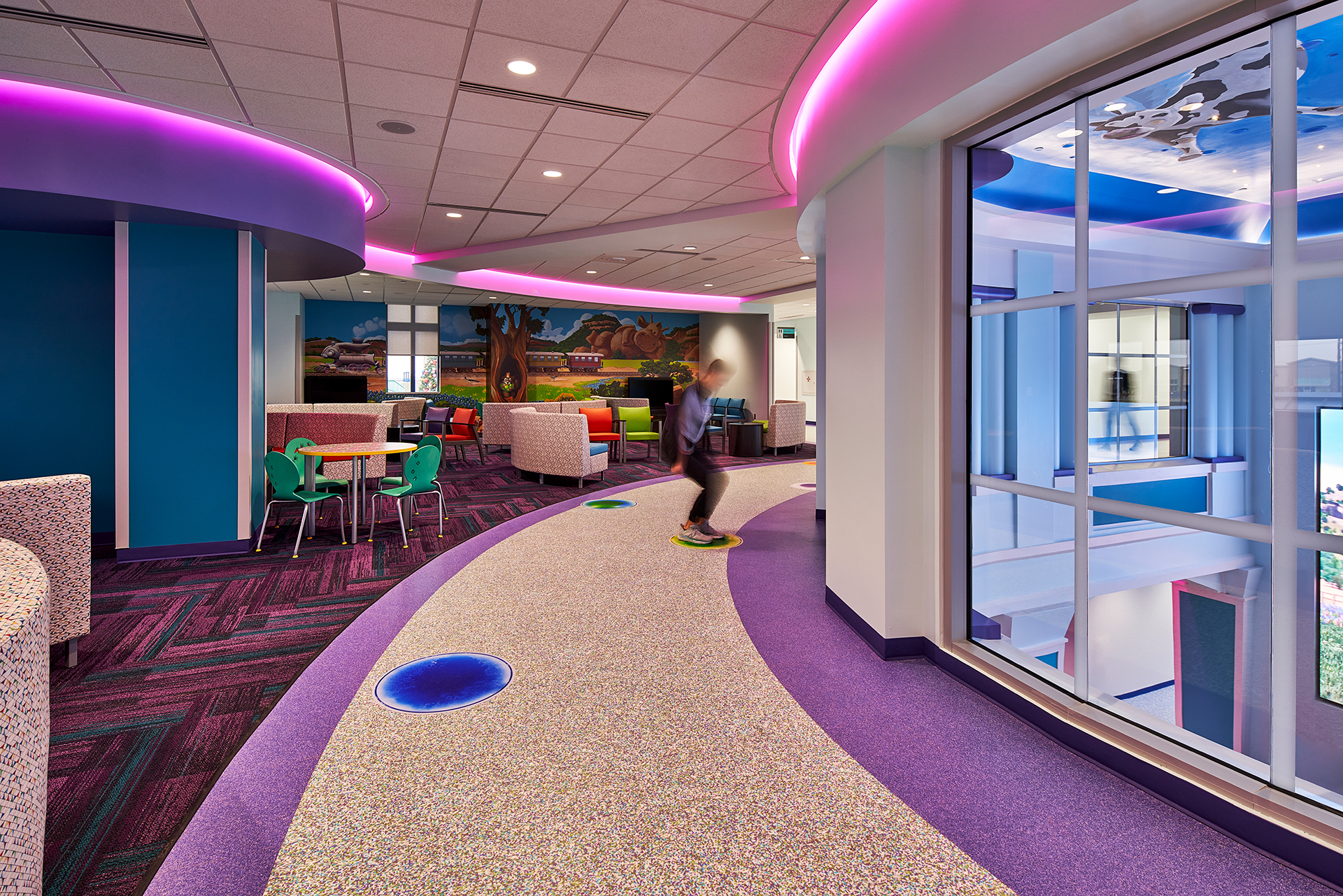 A colorful children's playroom with a boy playing, curved walkway, large windows, and vibrant purple and blue accents.