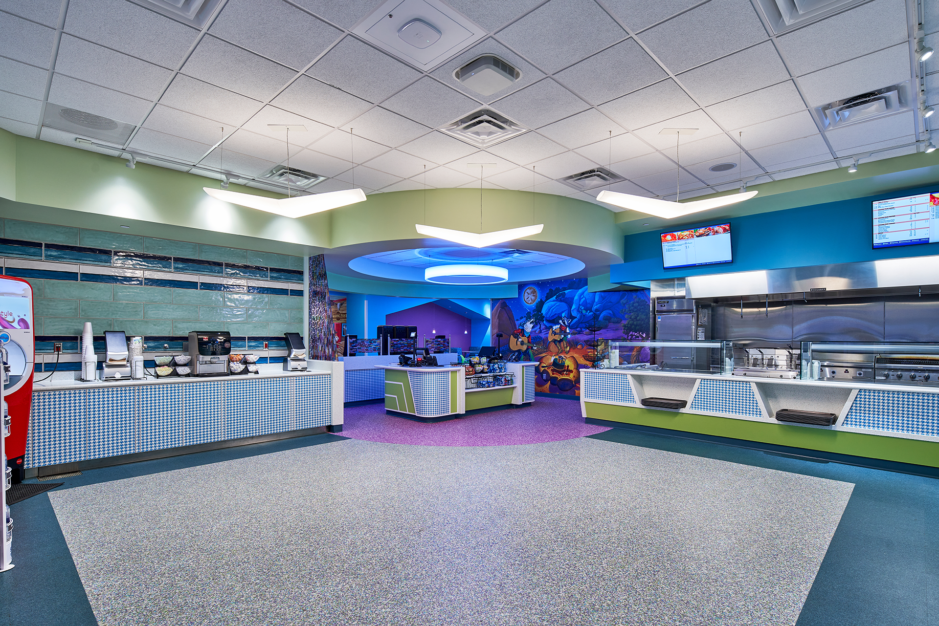 Interior of a brightly colored cafeteria with various food stations. There are blue, green, and purple accents, with a central, illuminated blue design.