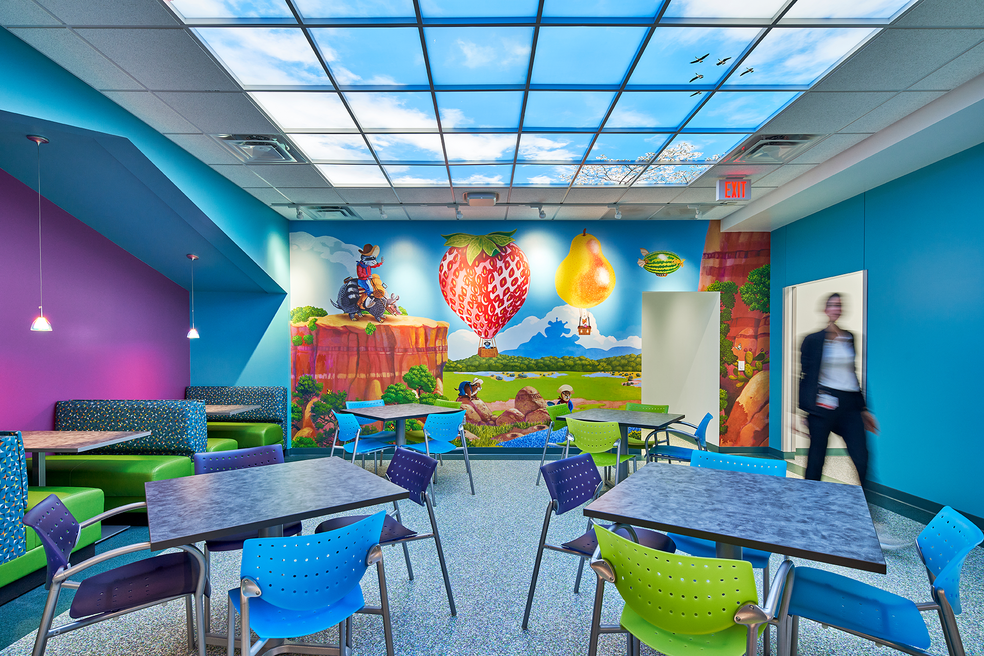 Colorful cafeteria with mural of hot air balloons and sky, tables with chairs, and person walking through a doorway.
