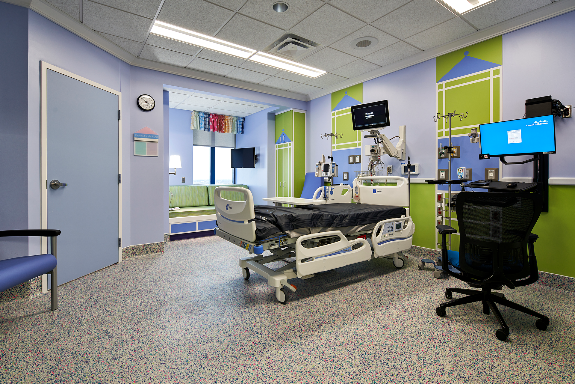 Hospital room with a bed, medical equipment, and a small seating area. Walls are blue and green with colorful accents.