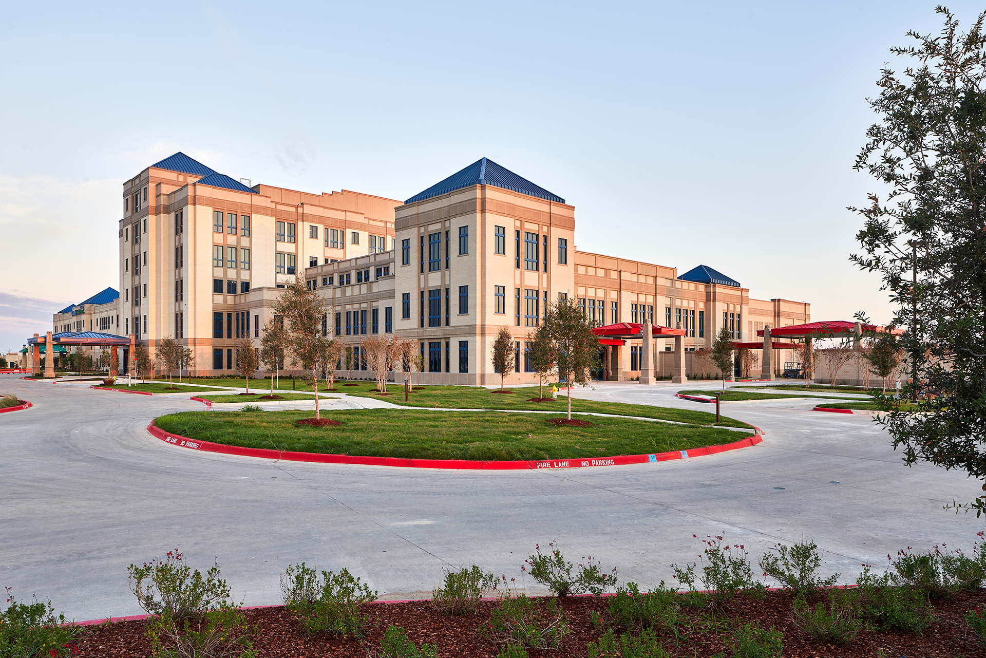 Large, multi-story building with light beige exterior, blue roof accents, and a circular driveway with a green center island.
