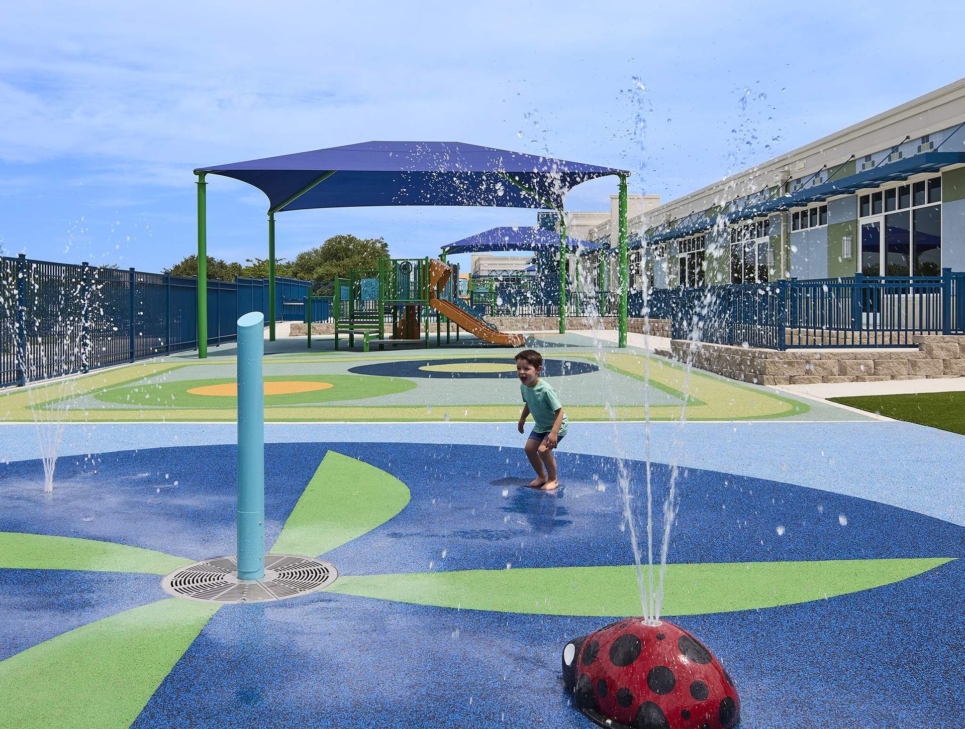 Child playing under water spray in a colorful playground with blue shade canopies and a ladybug ball