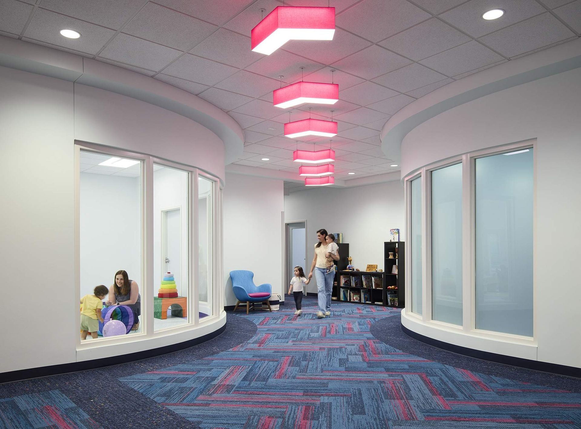 Curved hallway with pink ceiling lights, blue patterned carpet, and people sitting and standing near doorways