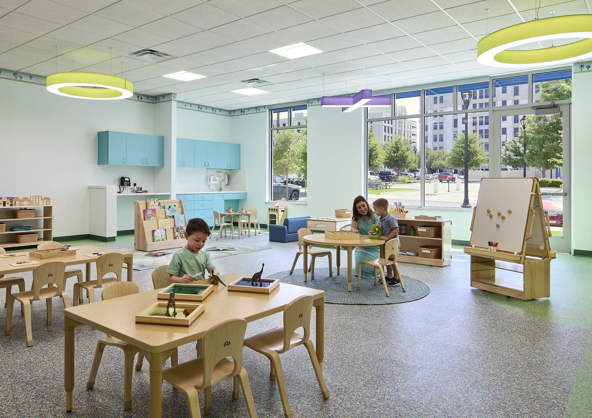 Bright classroom with children at tables, art easel, and large windows overlooking buildings outside