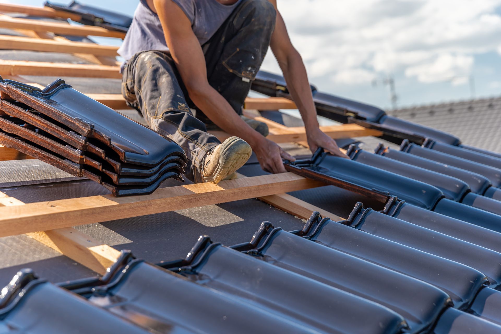 roofing contractor kneeling on a sunlit rooftop installing dark ceramic tiles along wooden battens