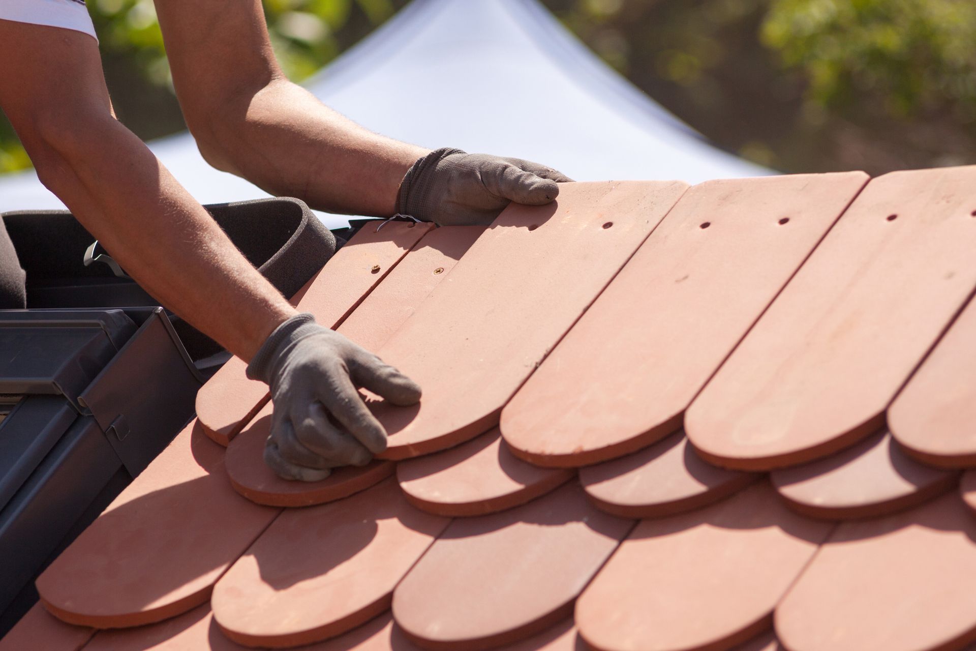 Male roofer with gray gloves lays a red tile on a roof, next to other tiles, under the sun.