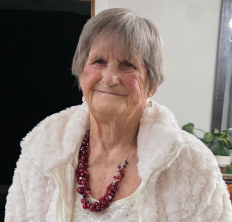 Smiling person in a white jacket and pink necklace, standing indoors near a plant.