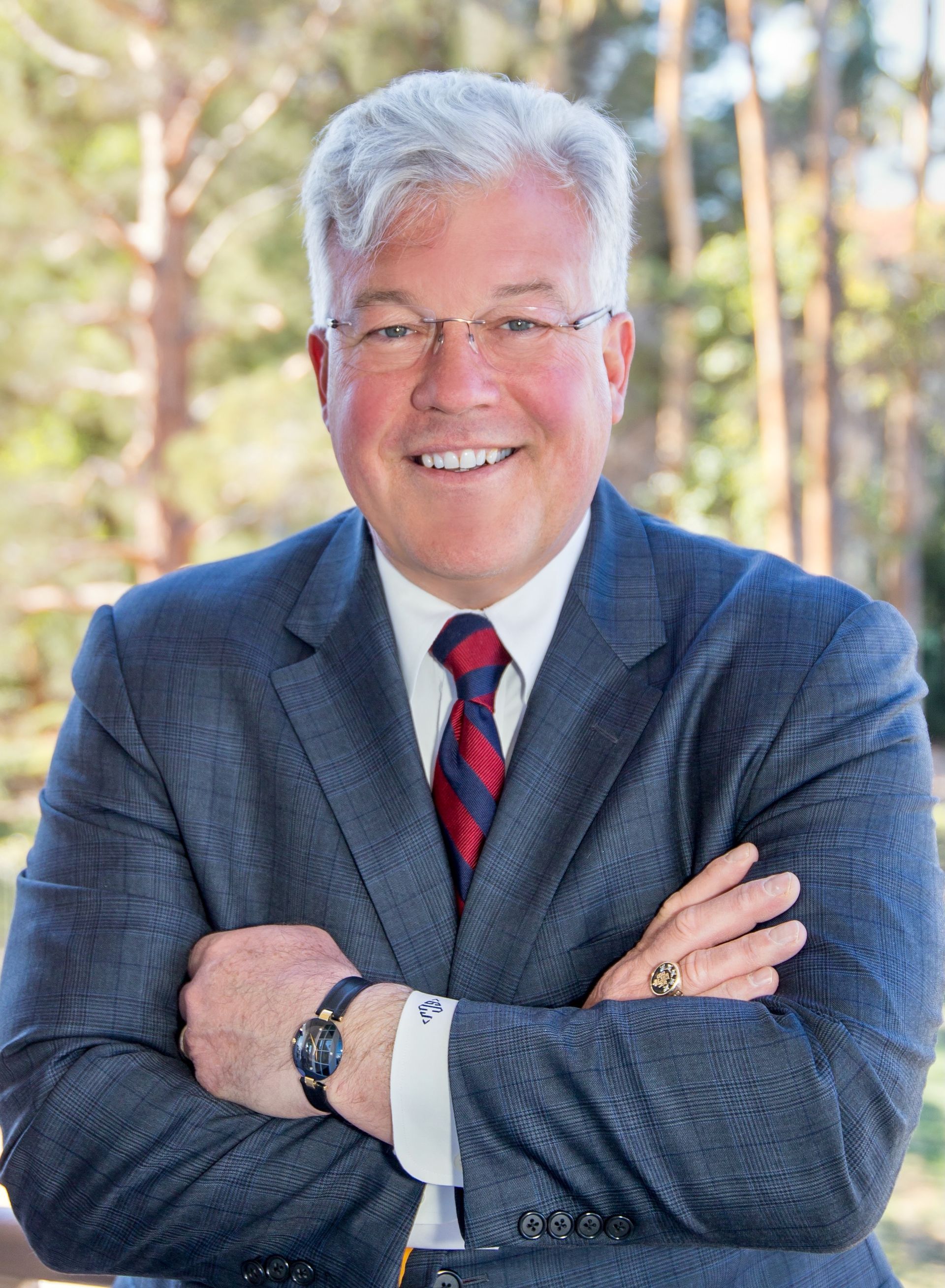 Man in suit with arms crossed smiling, in an outdoor setting.