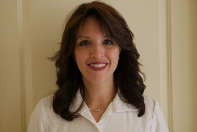 Woman with dark hair, smiling, wearing white shirt, in front of a light wall.