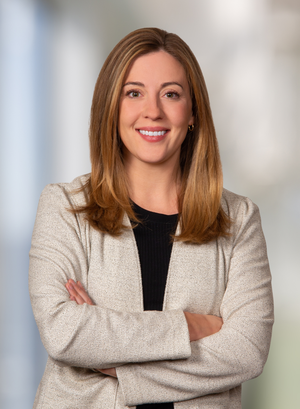 Woman with crossed arms, smiling, wearing blazer and black shirt, set against a blurred office background.