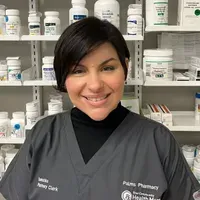 A woman in a pharmacy is smiling in front of a shelf full of pills.