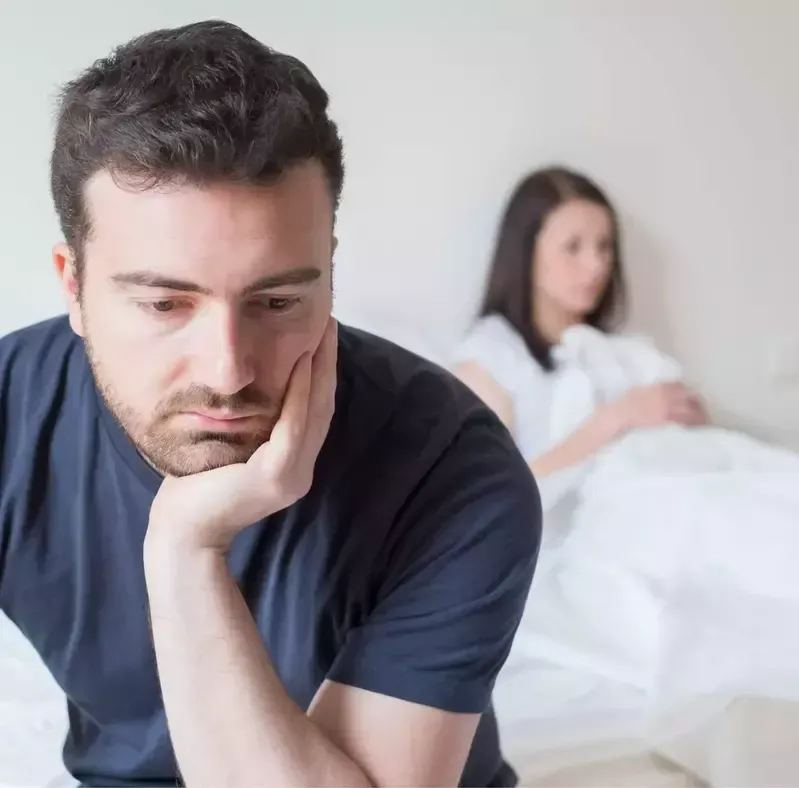 A man is sitting on a bed with his hand on his chin while a woman is laying in bed.