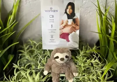 A stuffed sloth is sitting next to a woman holding a cup of coffee.