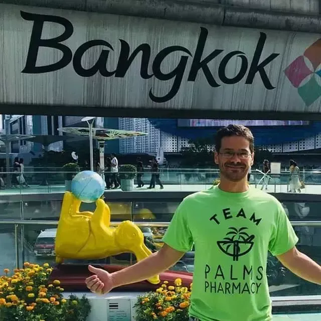 A man in a green team palms pharmacy shirt stands in front of a bangkok sign