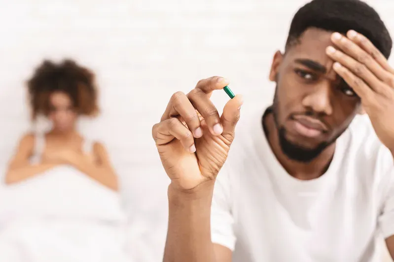 A man is holding a green pill in his hand while a woman is laying in bed.