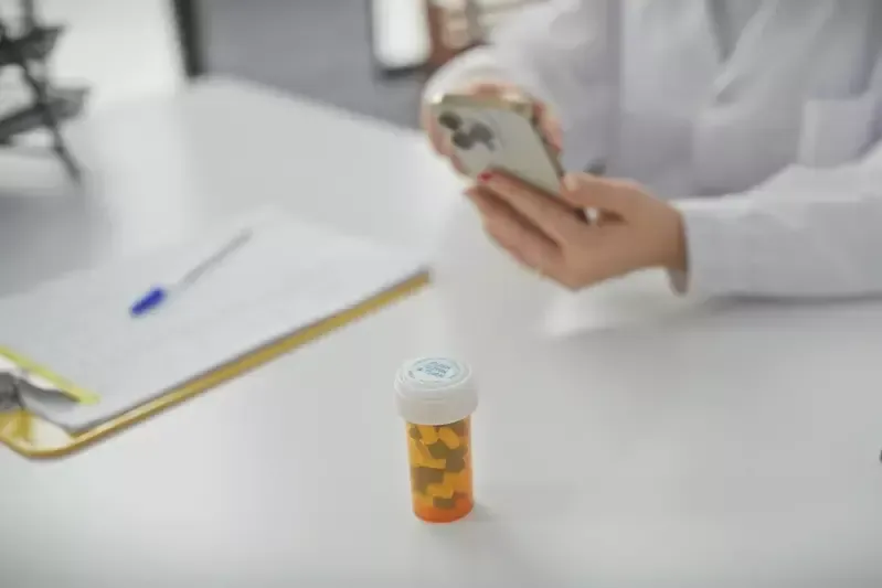 A doctor is sitting at a desk using a cell phone next to a bottle of pills.