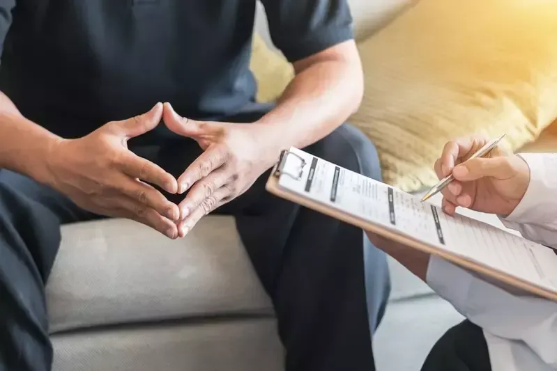 A man is sitting on a couch talking to a doctor who is holding a clipboard.