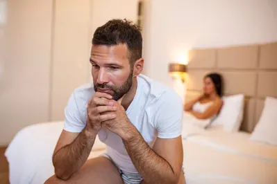 A man is sitting on the edge of a bed with a woman in the background.
