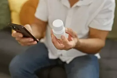 A man is sitting on a couch holding a bottle of pills and a cell phone.