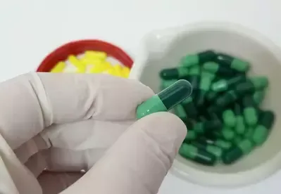 A person is holding a green capsule in front of a bowl of yellow pills.