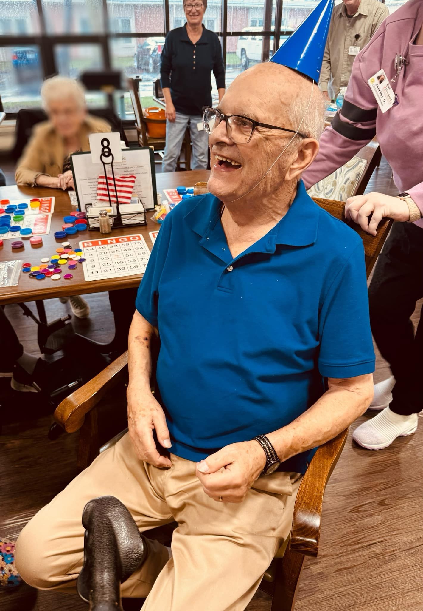 An elderly man wearing a blue shirt and a party hat is sitting in a chair.