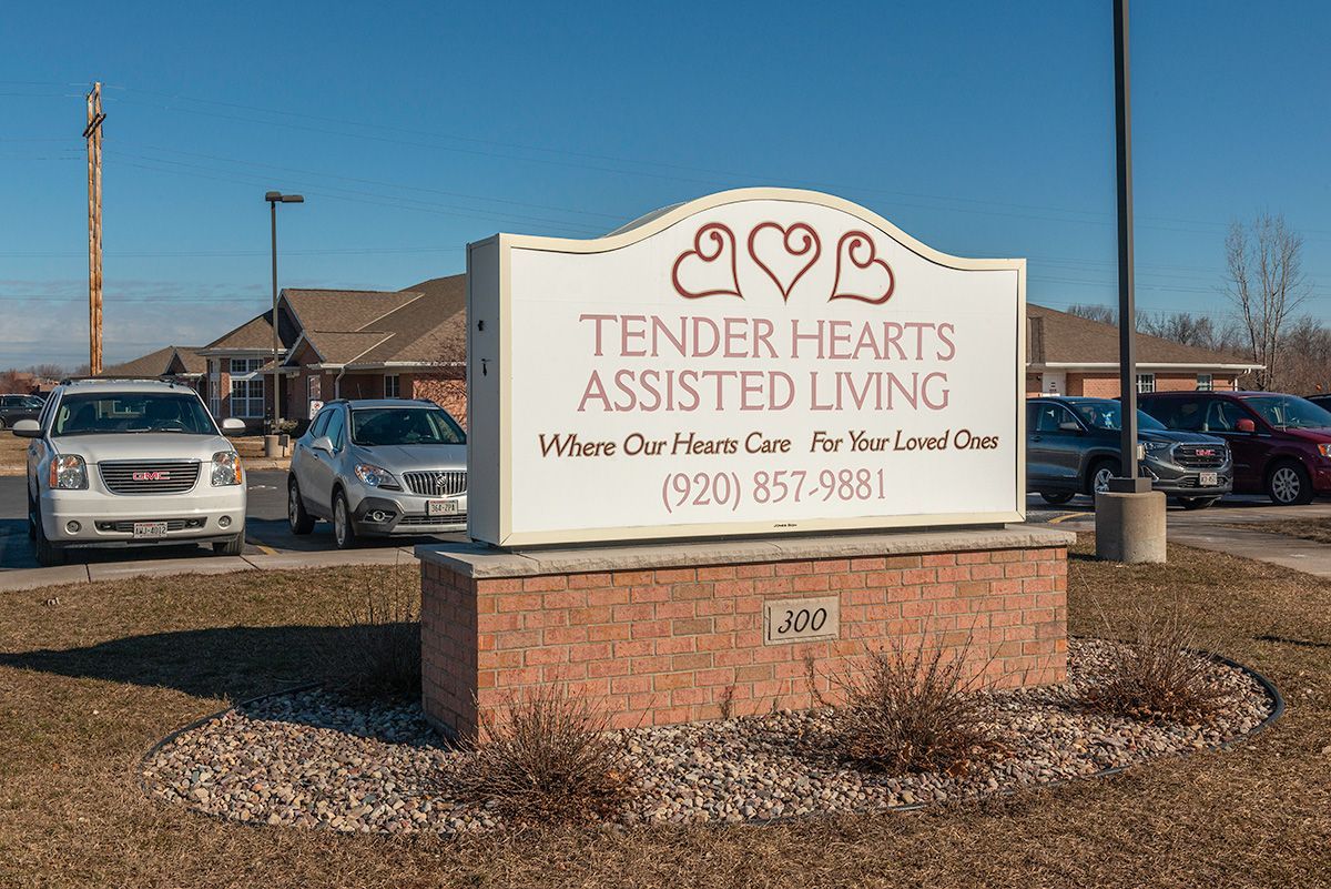 A sign for tender hearts assisted living is in a parking lot with cars parked in front of it.