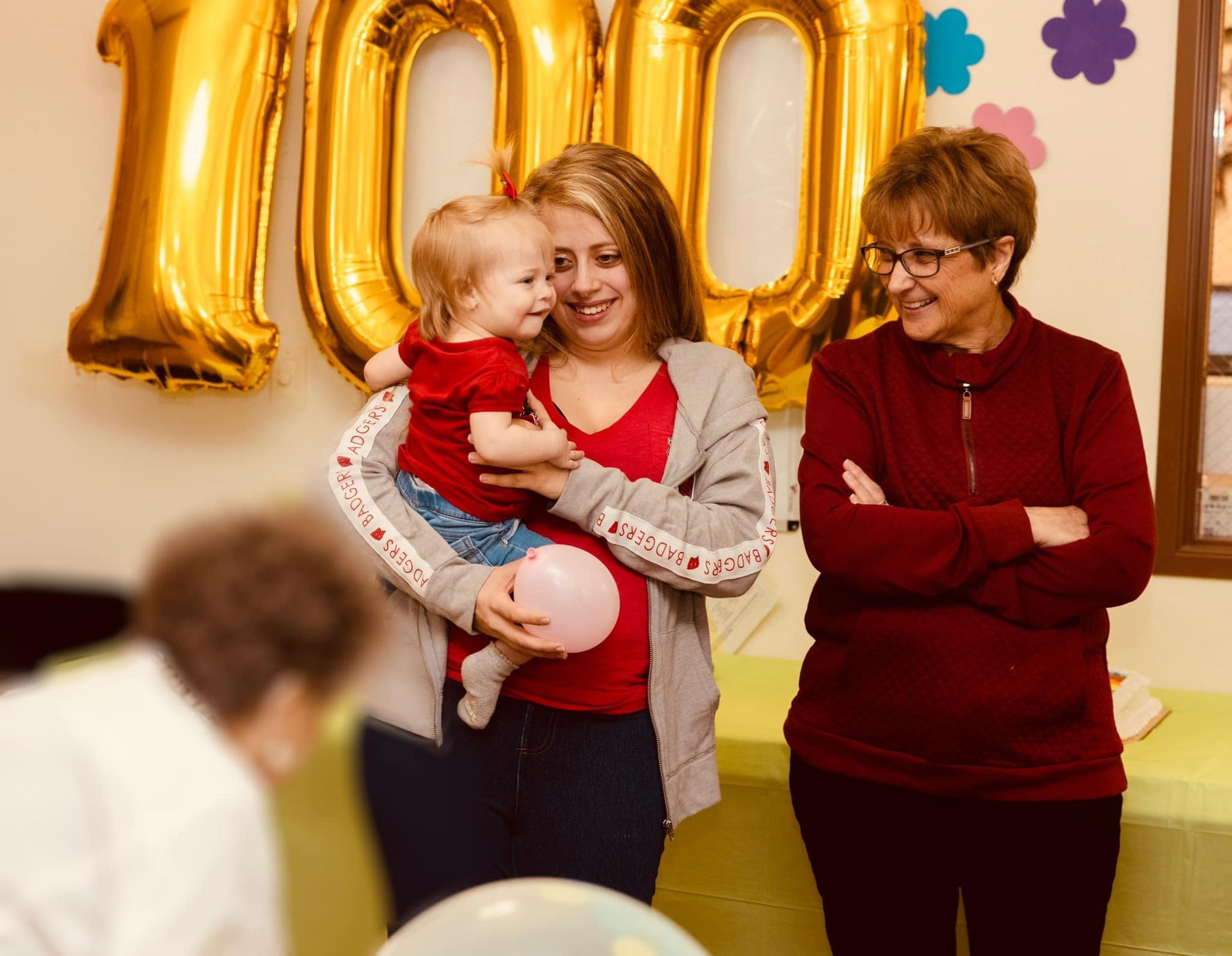 Two women are holding a baby in front of balloons that say 100