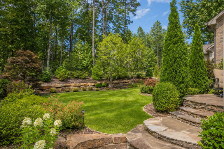 Landscaped backyard with green lawn, stone steps, shrubs, and tall trees under a blue sky