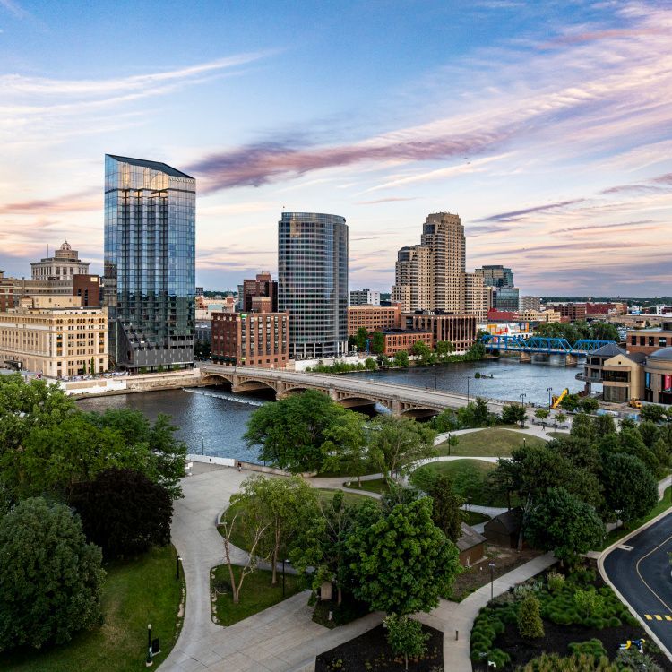 Downtown skyline with river, bridge, and park at sunset under a pastel sky