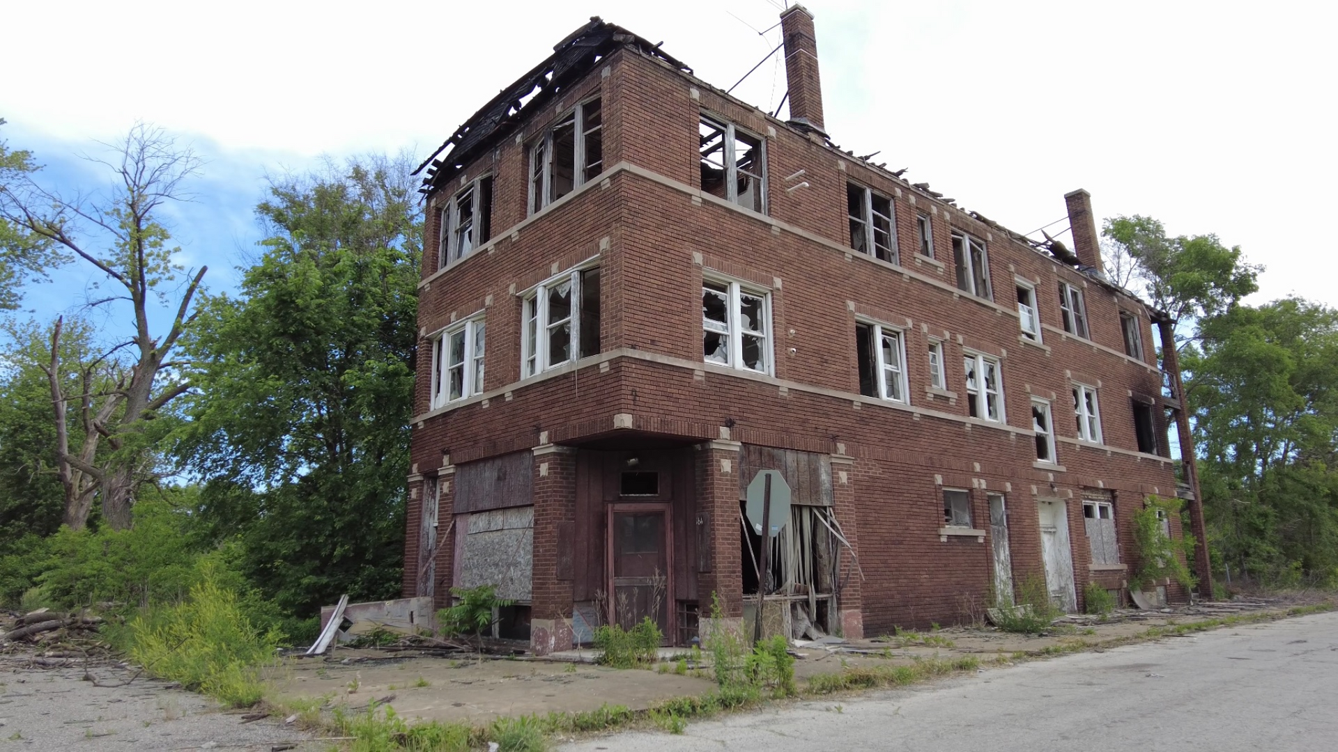 Abandoned red-brick building with broken windows and overgrown weeds beside a road