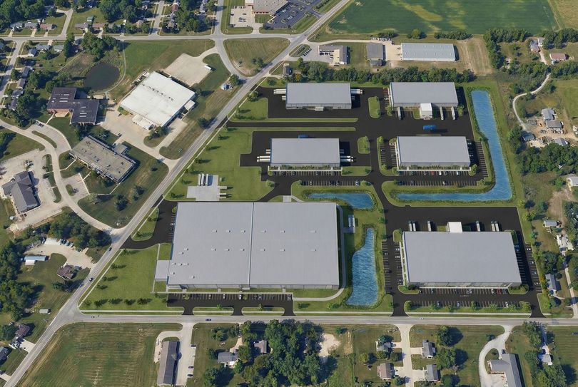 Aerial view of an industrial complex with large gray warehouses, roads, parking lots, and surrounding greenery.