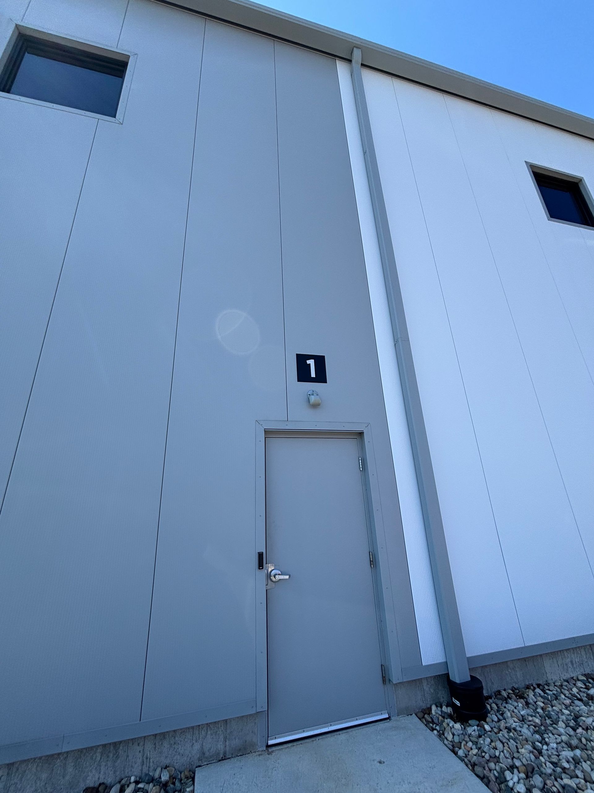 Gray industrial building exterior with a metal door, number 1 sign, and gravel ground.