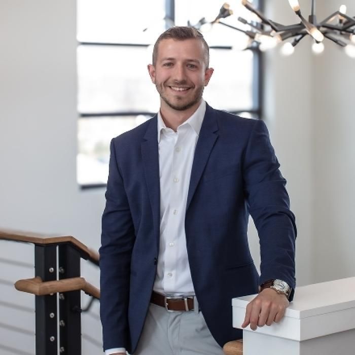 Man in a navy suit standing by a staircase in a bright office, smiling with one hand on a white railing