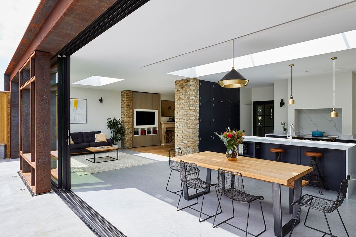 Open-air modern kitchen and dining area with wood table, black stools, and retractable glass wall.