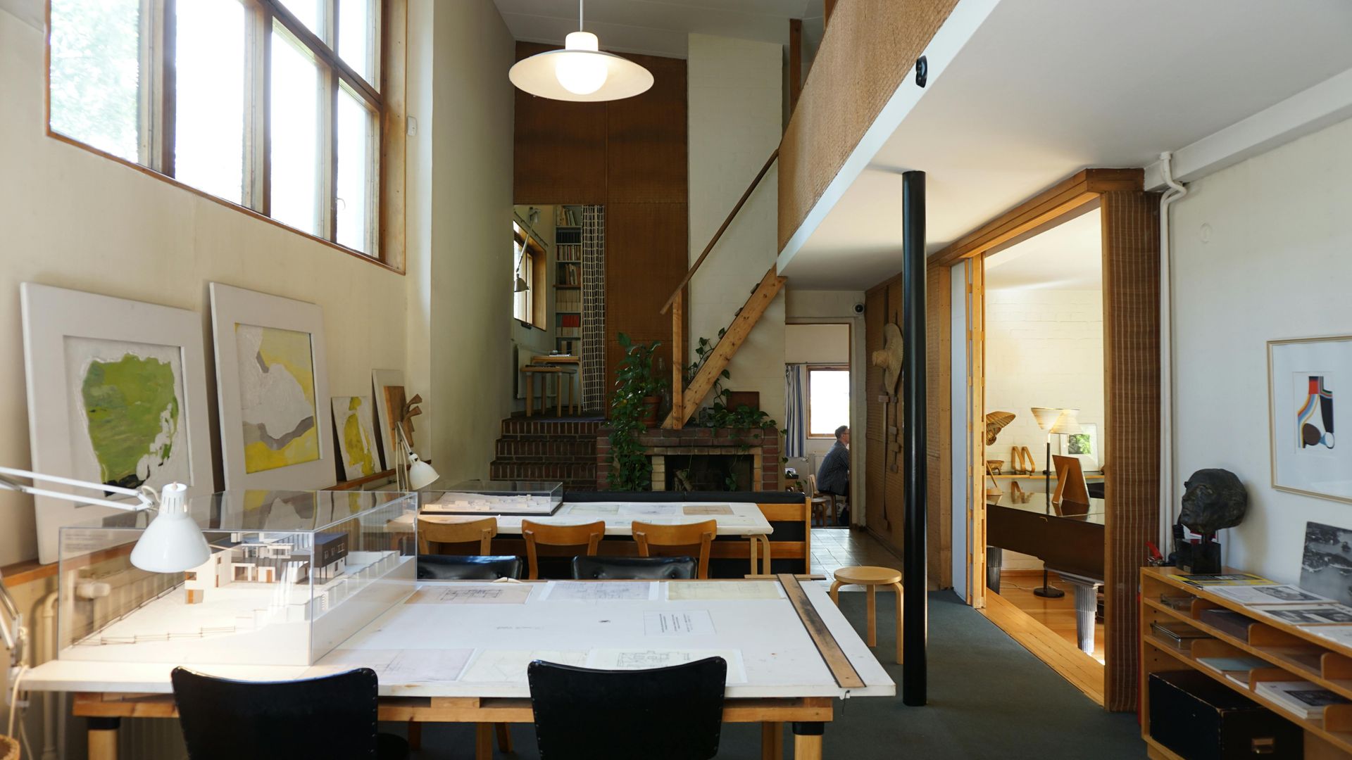 Bright loft office with large worktables, computers, shelves, and tall windows.