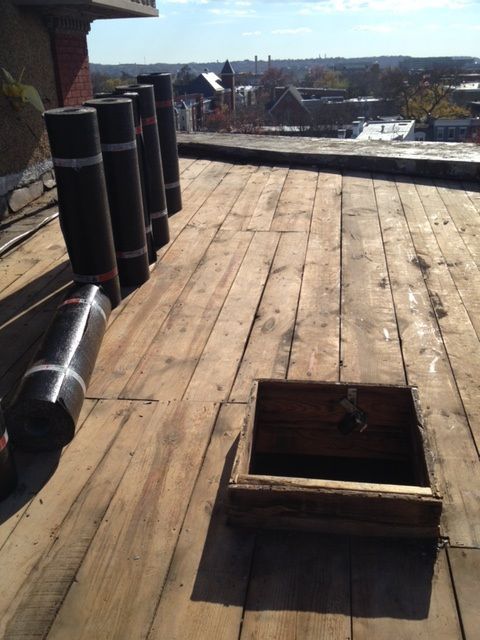 Flat rooftop with wooden decking, black vent pipes, and an open hatch under a bright sky.
