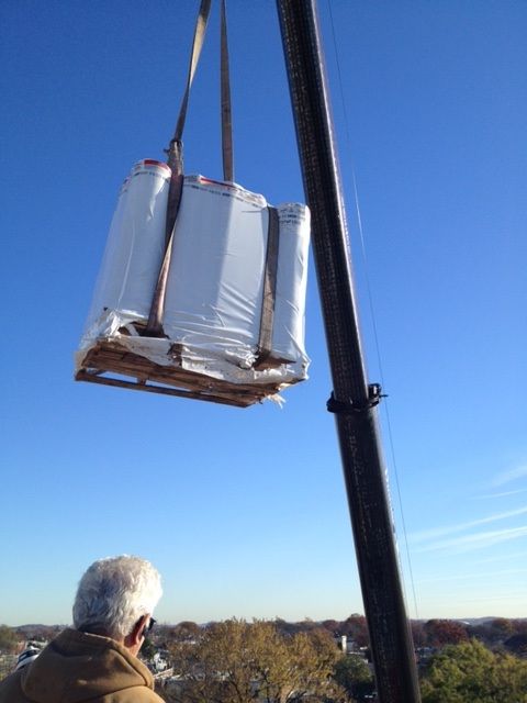 Crane lifting a wrapped pallet high above a tree-lined area under a clear blue sky