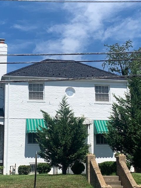 White two-story house with green awnings, central evergreen tree, and blue sky overhead