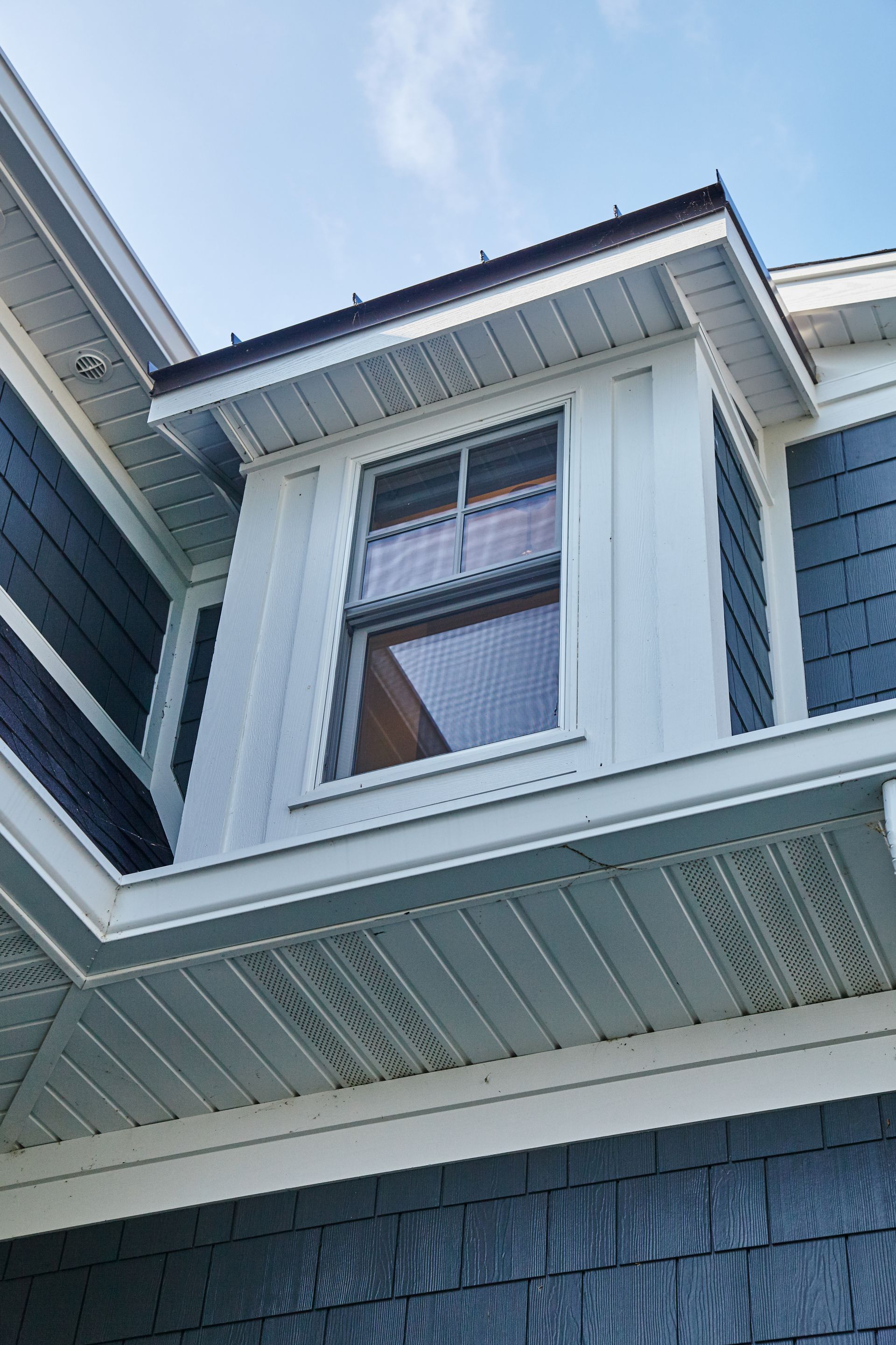 Close-up of a house exterior with a white-trimmed window and gray shingle siding under the roof eaves