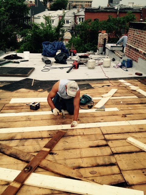 Worker kneeling on a rooftop, measuring wooden planks during roof repair.