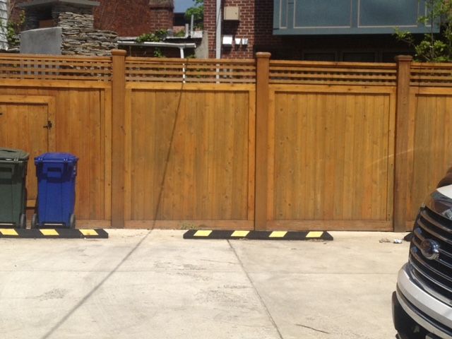 Wooden fence with gate, blue trash bin, and parked car at the edge of a driveway.