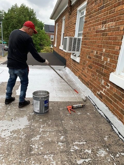 Worker painting exterior brick wall beside walkway, with bucket and roller on the ground.