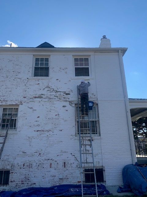 Worker on a ladder painting a white house exterior with peeling paint under a blue sky