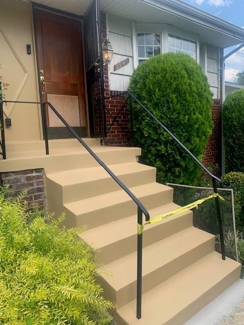 Front steps to a house with beige stairs, black railings, and green shrubs by the entrance